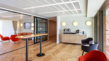 A modern interior with a wooden herringbone floor, a central table, red chairs, a kitchen area with a coffee machine, and a glass partition.
