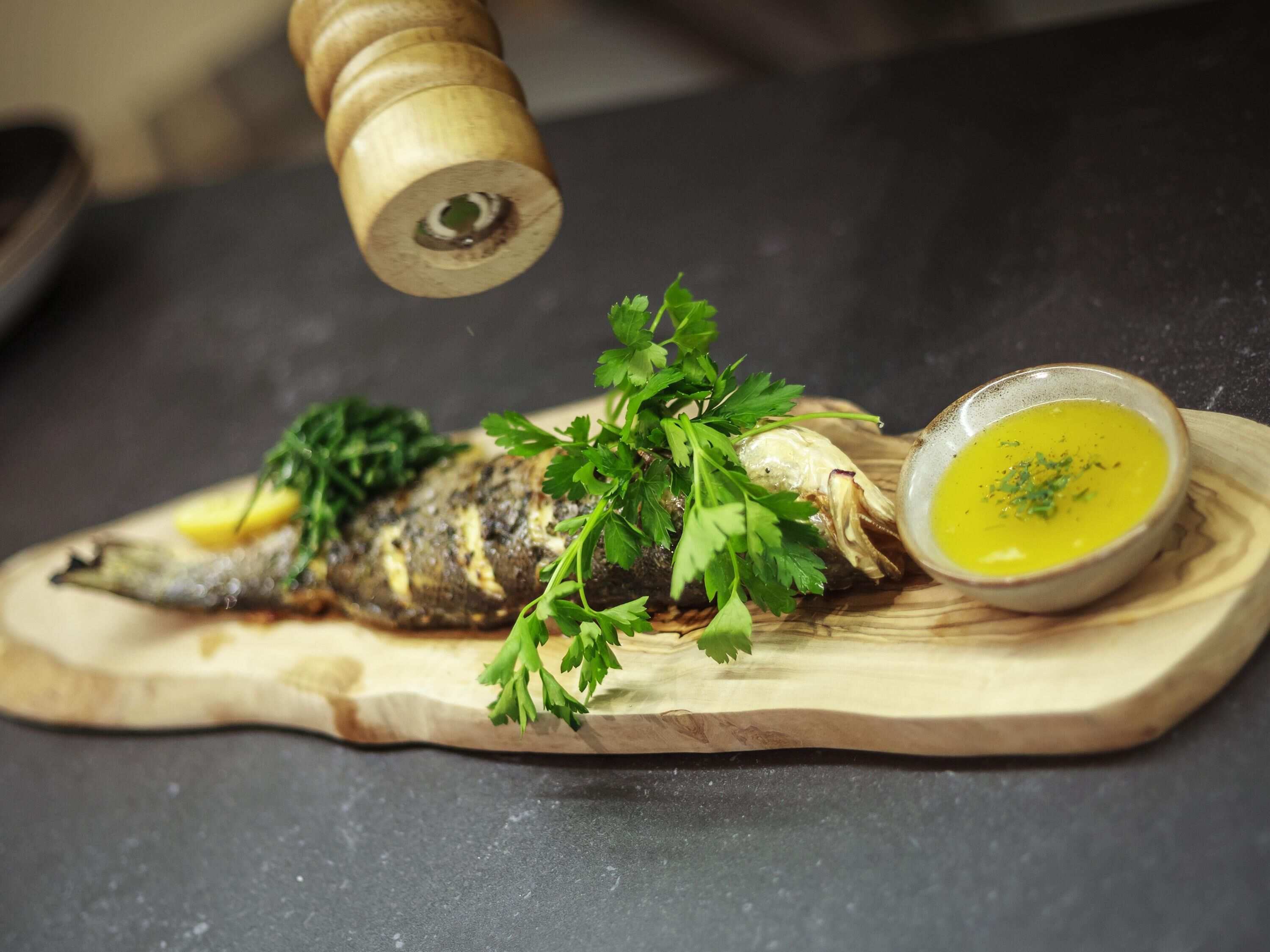 A grilled fish on a wooden board with a bowl of sauce and parsley garnish.
