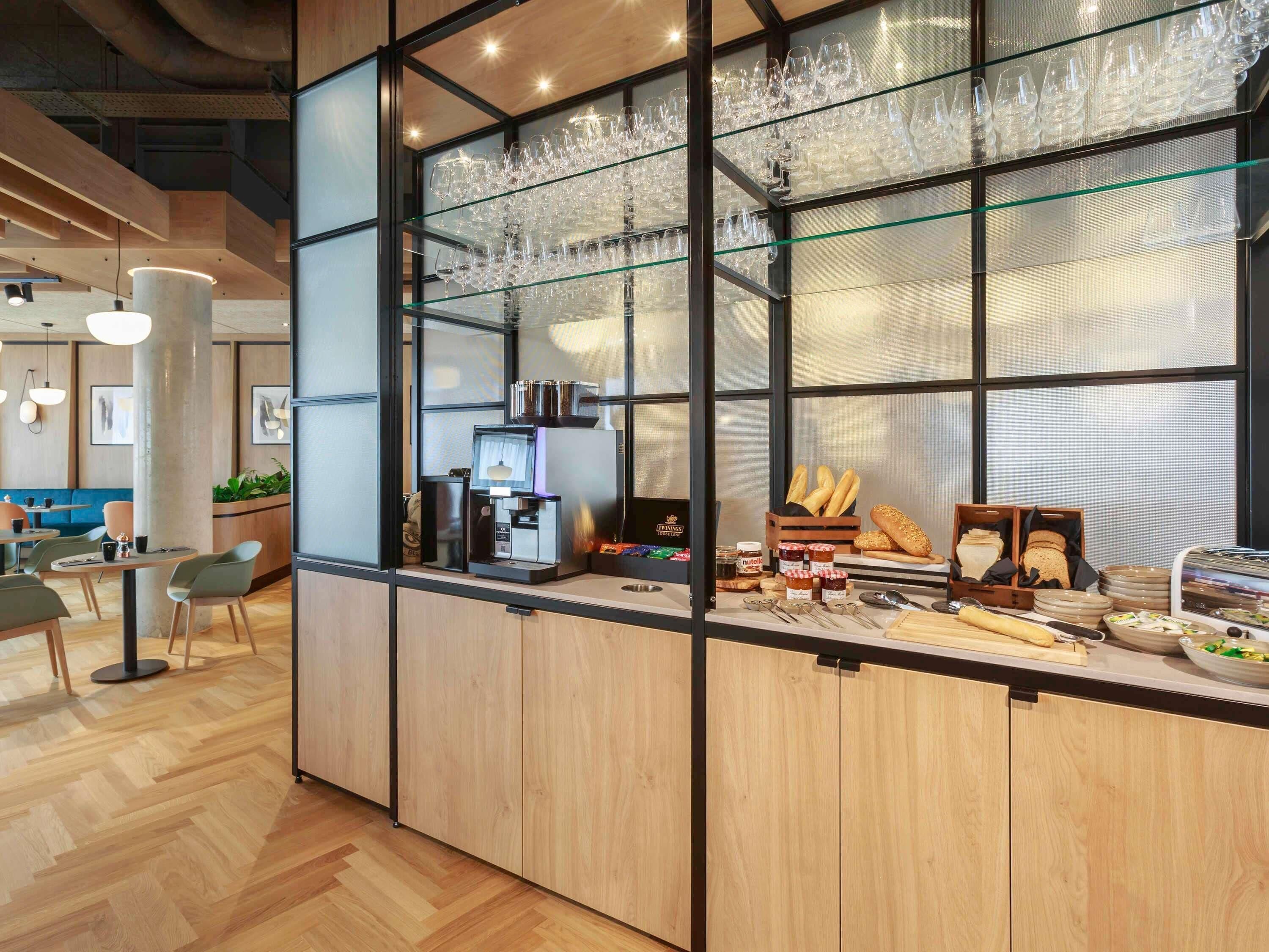 A modern cafe with a wooden counter, glass display of wine glasses, coffee machines, and a variety of pastries.