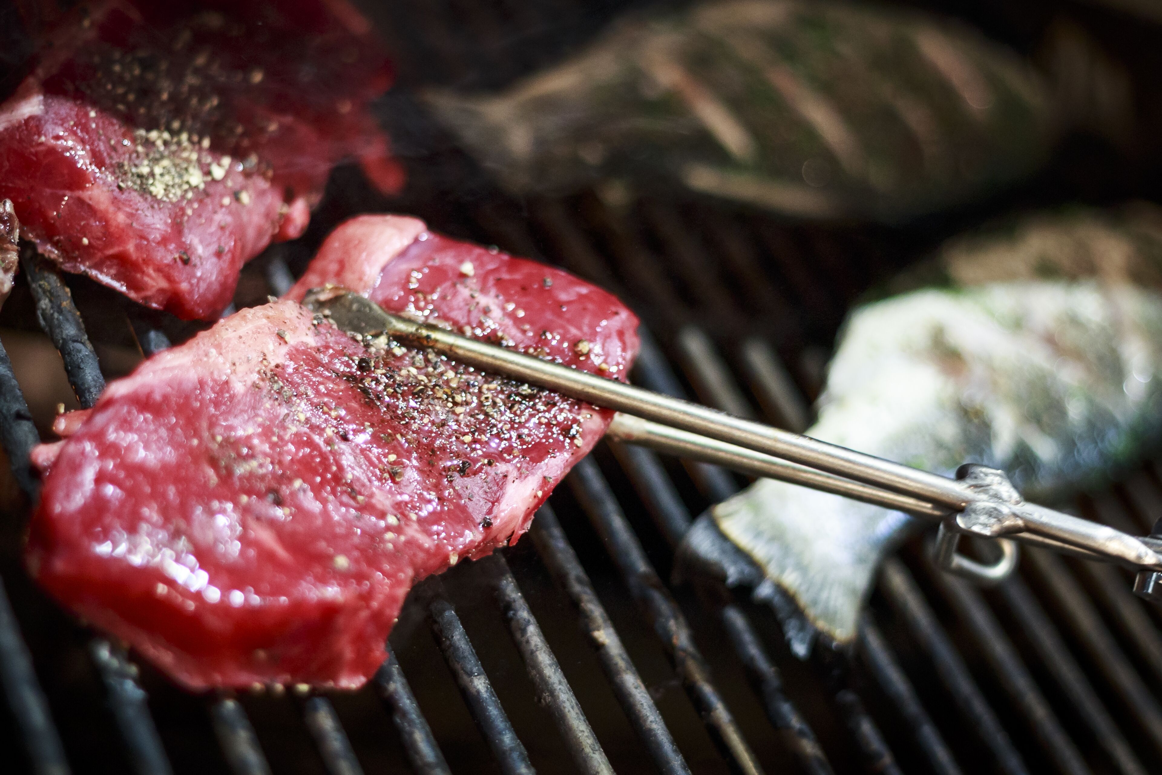 Steaks on a grill with a tongs resting on top.