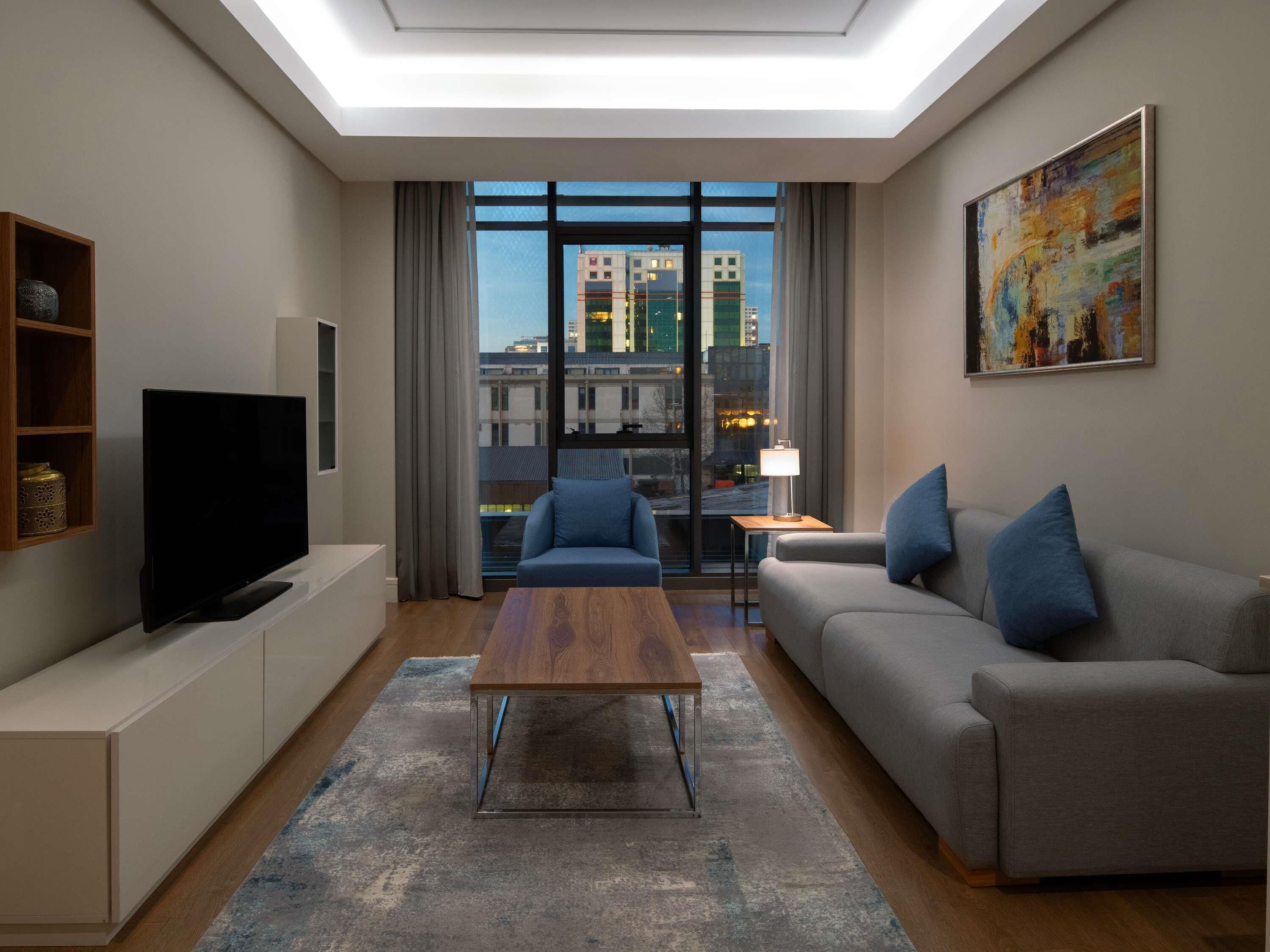 A modern living room with a flat-screen TV, a wooden coffee table, a grey sofa with blue cushions, and a city view through large windows.