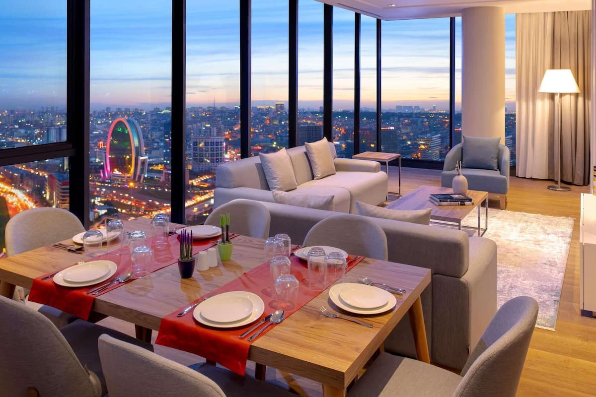 A high-rise hotel room with a dining area, a sofa, and a view of a cityscape at dusk.