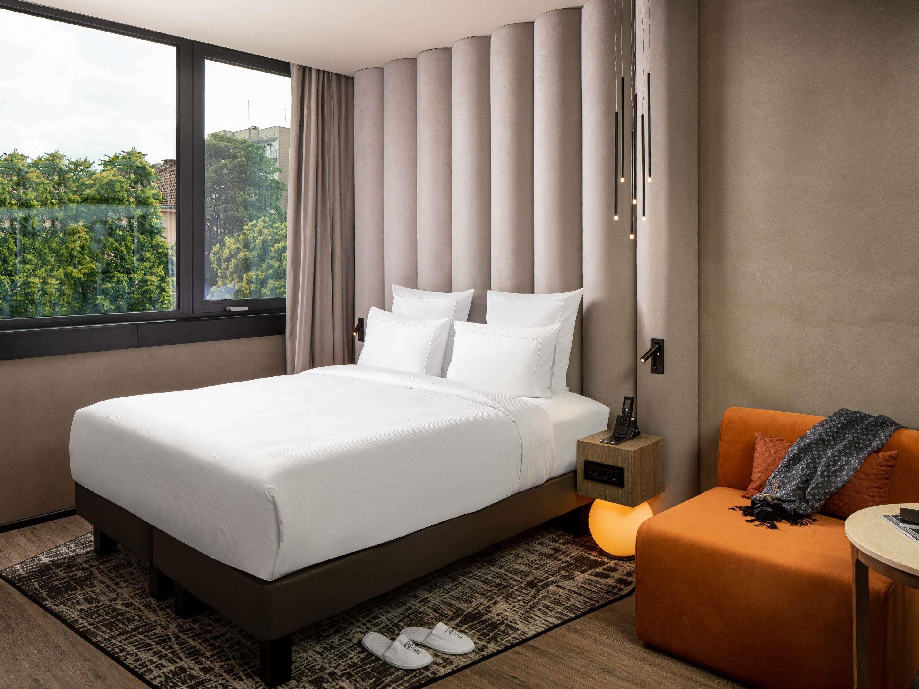 Una habitación de hotel moderna con una cama grande, una mesita de noche con teléfono, una pequeña zona de estar naranja y una ventana con vistas a zonas verdes.
