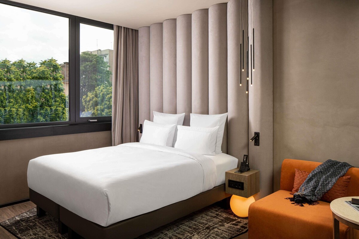 Une chambre d’hôtel moderne avec un grand lit, une table de chevet avec un téléphone, un petit coin salon orange et une fenêtre donnant sur un paysage verdoyant.