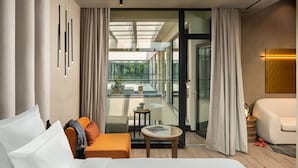 Une chambre d’hôtel moderne avec un grand lit, un canapé, une petite table et une vue sur un balcon extérieur, accessible par une porte vitrée.