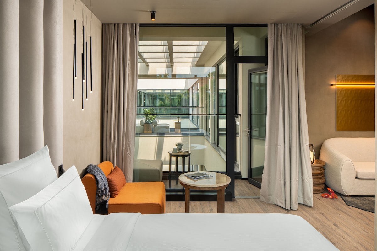 Ein modernes Hotelzimmer mit einem großen Bett, einer Couch, einem kleinen Tisch und Blick durch eine Glastür auf einen Außenbalkon.
