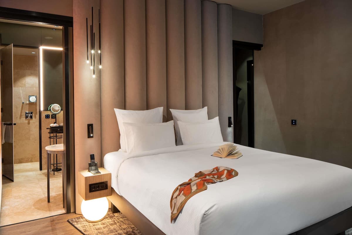 Habitación de hotel moderna con una cama grande, una mesita de noche con lámpara, un espejo y un baño visible a través de una puerta de cristal.