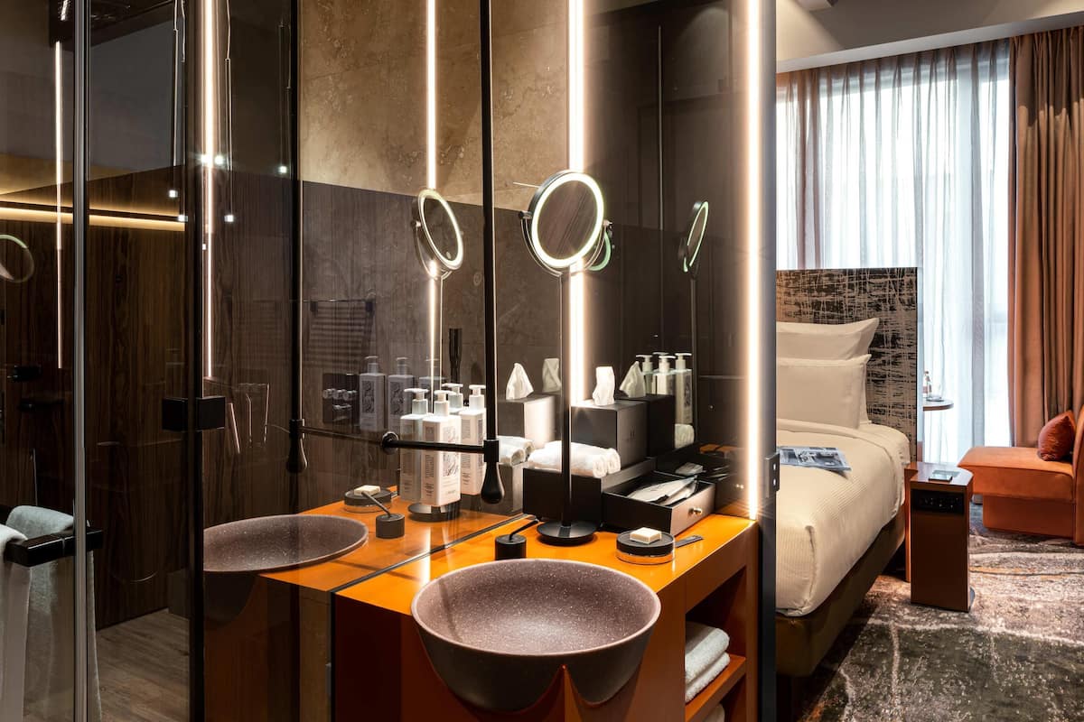 Una habitación de hotel moderna con una cama grande, un tocador con dos lavamanos, un espejo con luces y vistas a la ciudad a través de amplias ventanas.