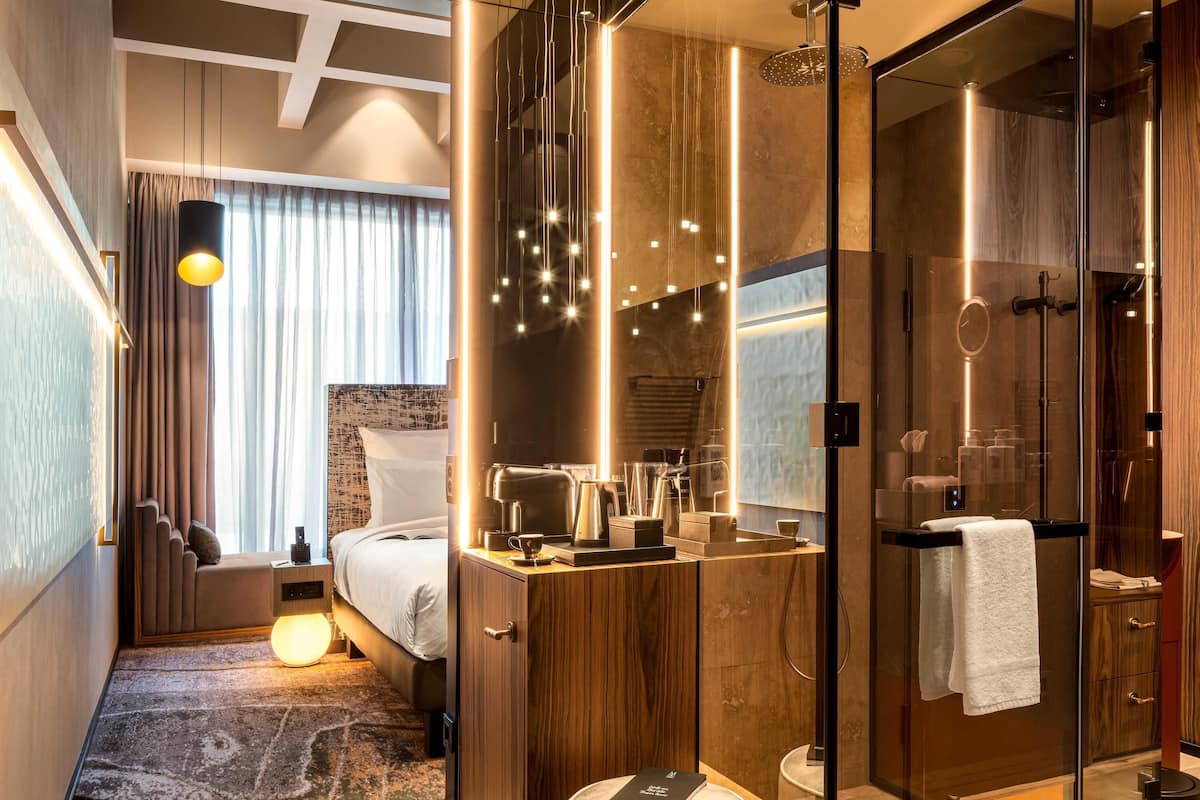 Una habitación de hotel moderna con una cama, mesita de noche, lámpara y un espejo grande.