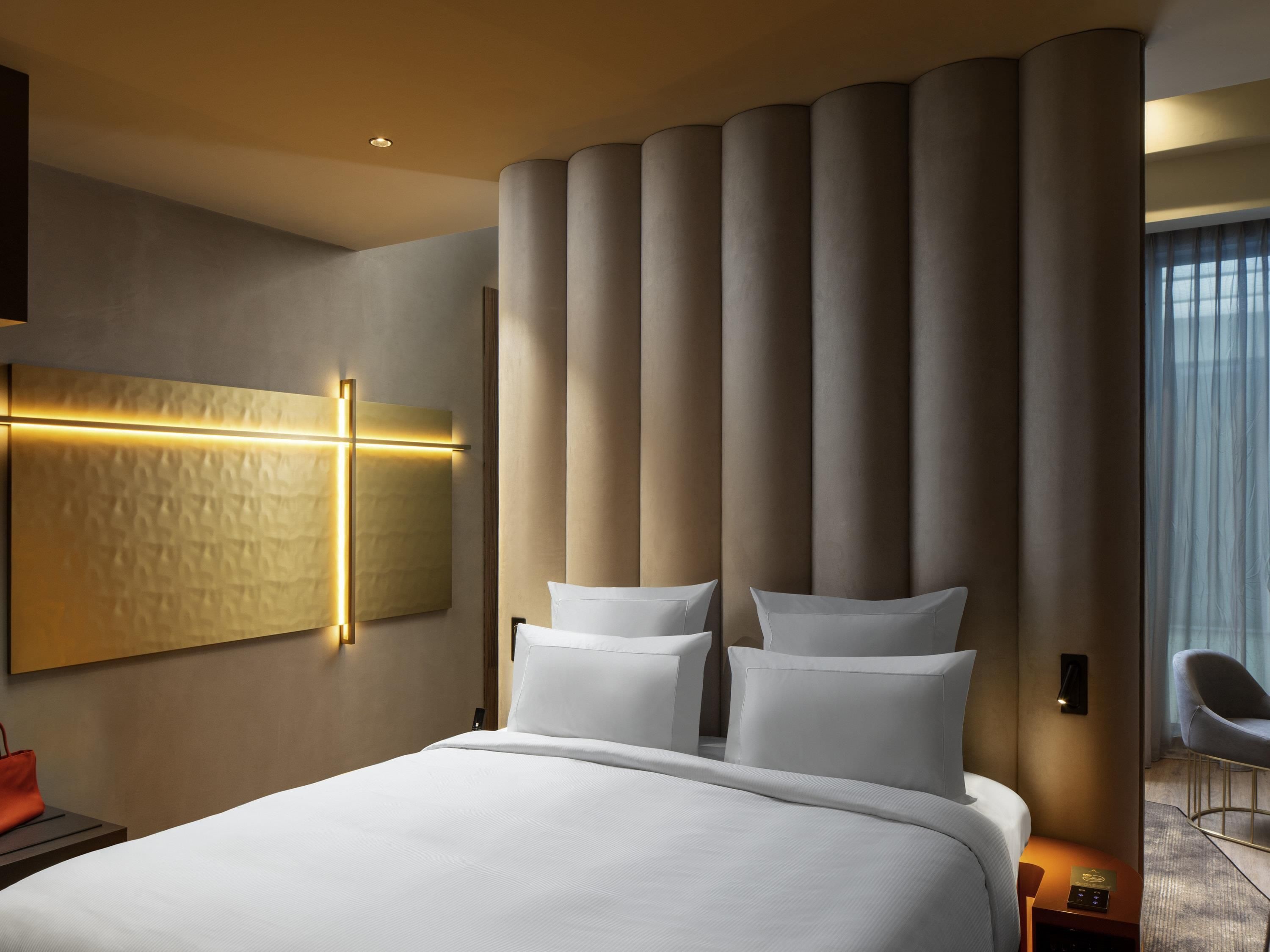 Quarto de hotel moderno com uma cama grande, luminária fixada na parede e uma janela ampla com cortinas.