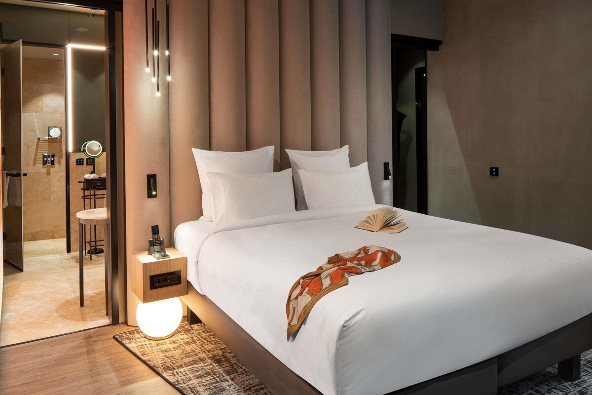 Une chambre d’hôtel moderne avec un grand lit, une table de chevet avec une lampe, un miroir et une salle de bain visible à travers une porte en verre.