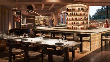 Interior de restaurante con varias mesas, sillas y una zona de bar central con estanterías llenas de botellas.