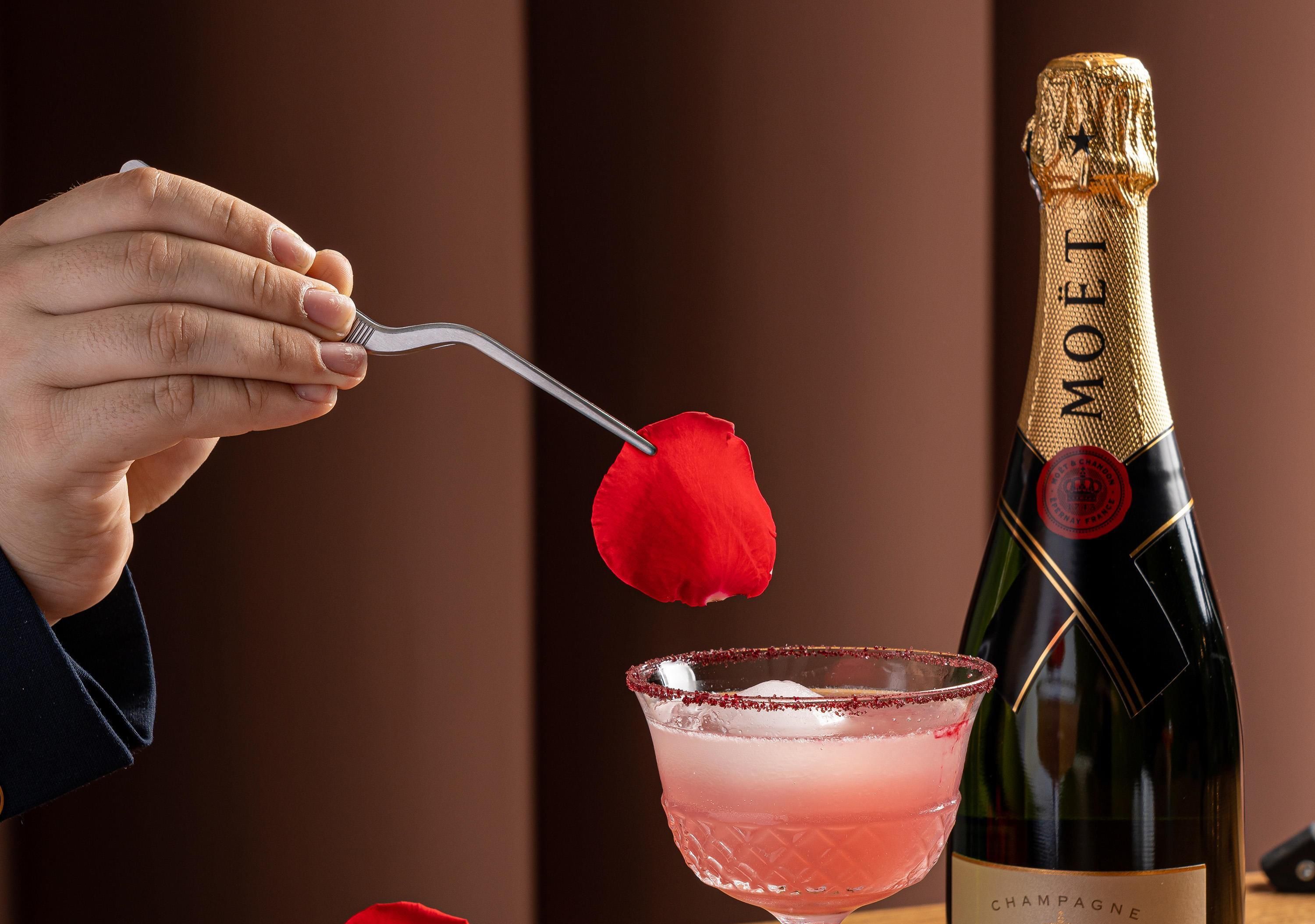 Una mano sosteniendo un tenedor con una rosa, una botella de Moët champagne y un cóctel rosado con hielo.