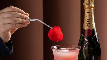 Una mano sosteniendo un tenedor con una rosa, una botella de Moët champagne y un cóctel rosado con hielo.