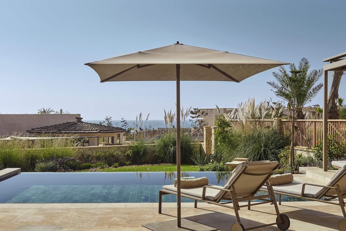 Zona de piscina con sillones de descanso y una sombrilla, rodeada de vegetación y vista al mar.