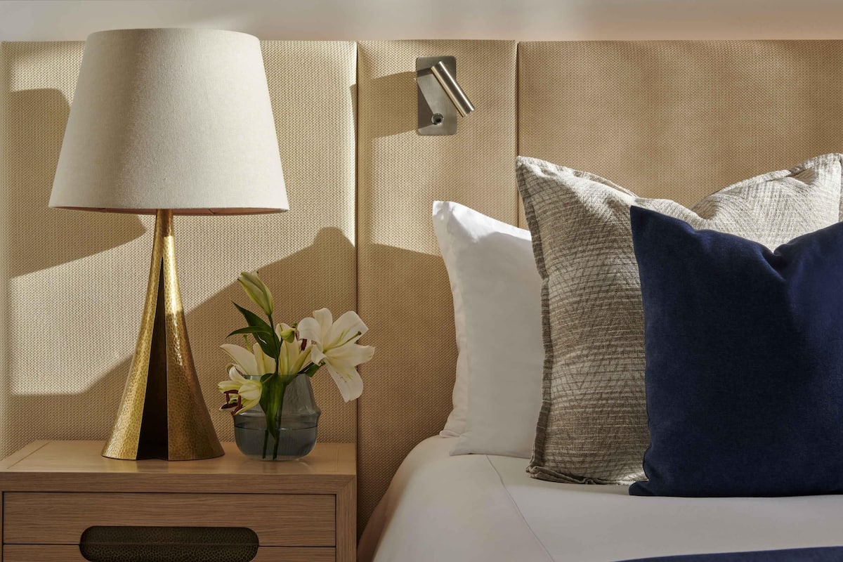 Una cama bien hecha con almohadas blancas y azules, una lámpara de noche y un pequeño florero con flores.