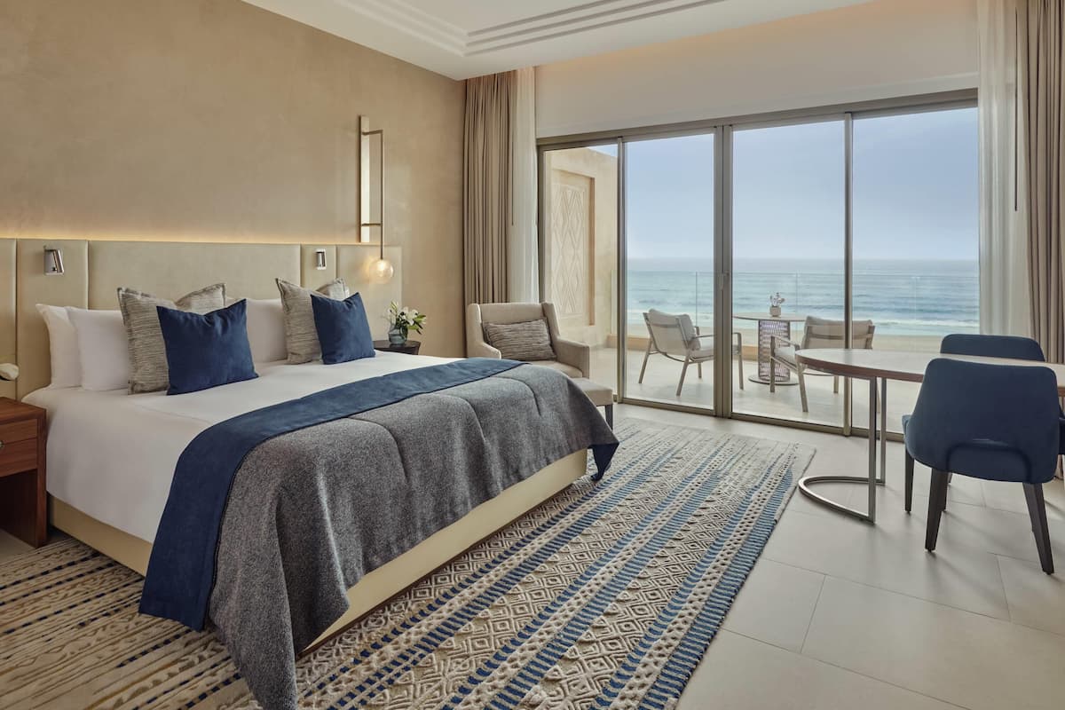 Habitación de hotel con una cama grande, un balcón con vista al océano, un comedor y un diseño moderno.