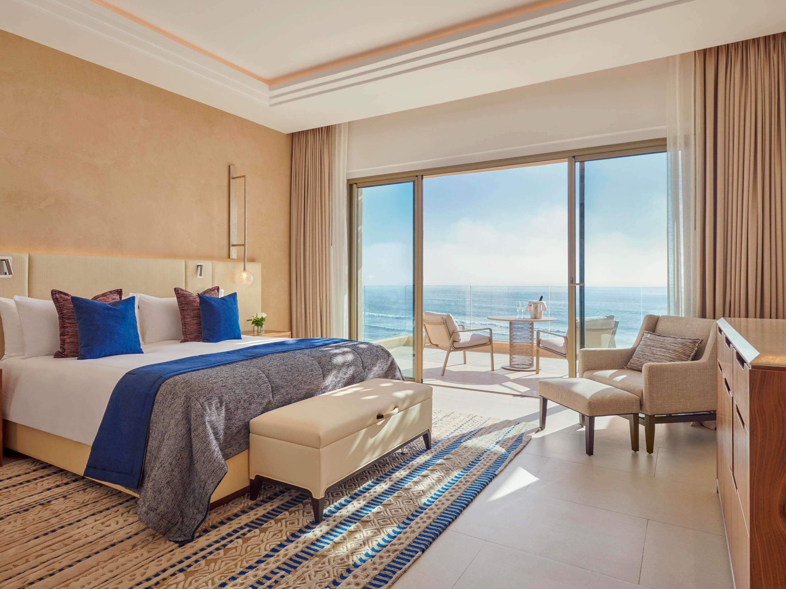 A hotel room with a large bed, a sitting area with a view of the ocean, and a balcony with a table and chairs.