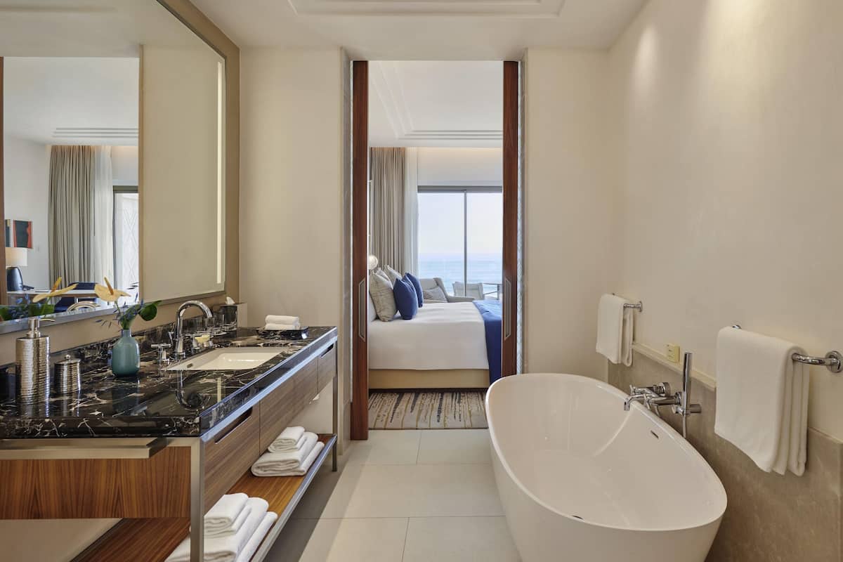 Un baño de hotel moderno con una bañera grande, un tocador con encimera de mármol, un espejo y vistas a un balcón con cama.