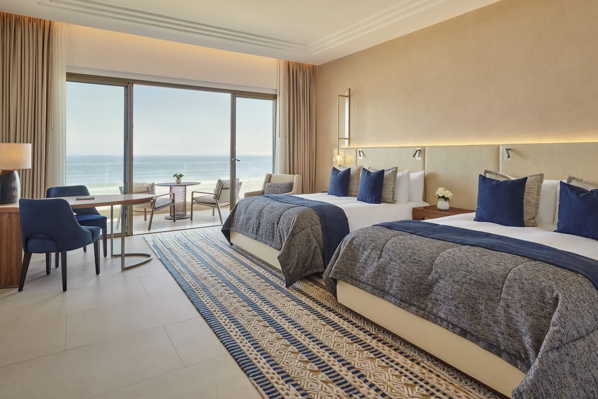Habitación de hotel con dos camas, un escritorio, una silla, una mesita y vista al océano.
