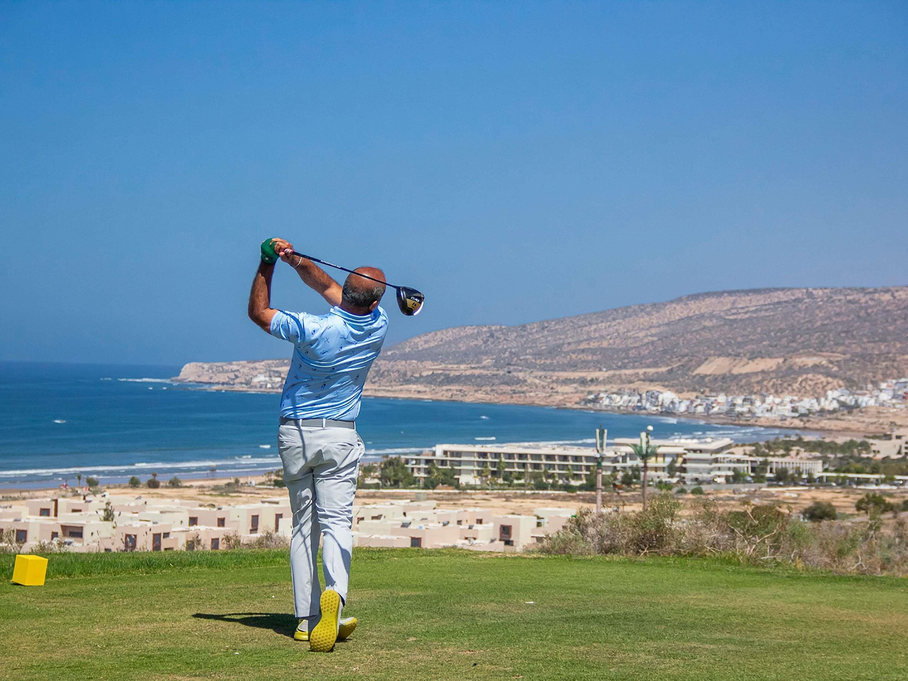 Un homme joue au golf par une journée ensoleillée, avec une vue dégagée sur l’océan et un paysage montagneux en arrière-plan.