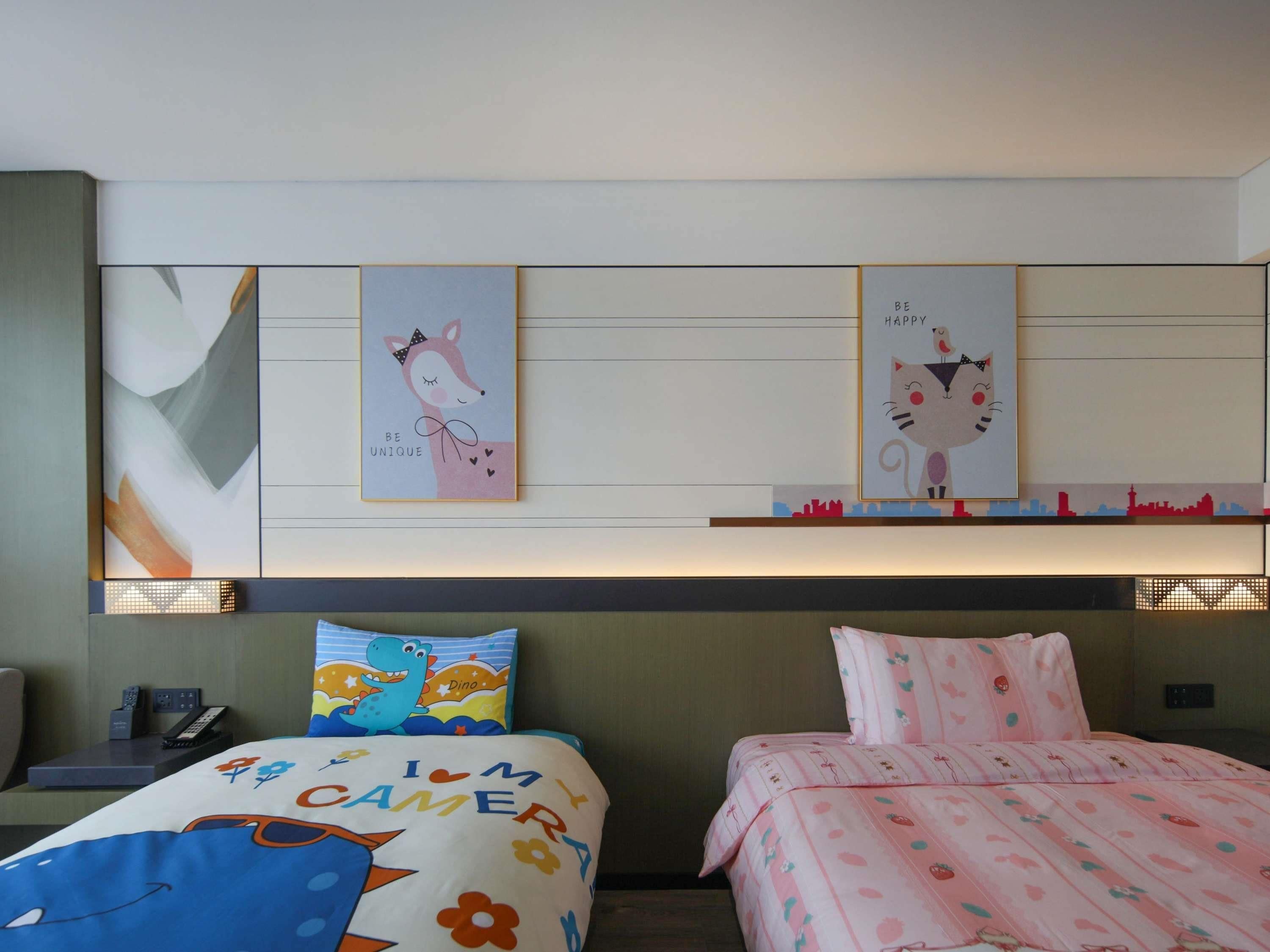 A hotel room with two beds, each with a distinct design. The left bed has a blue and orange pattern with the text "I'M CAMERA" and the right bed has a pink and white pattern with strawberries. Above the beds, there are two framed pictures: one of a fox with the text "BE UNIQUE" and another of a cat with the text "BE HAPPY".