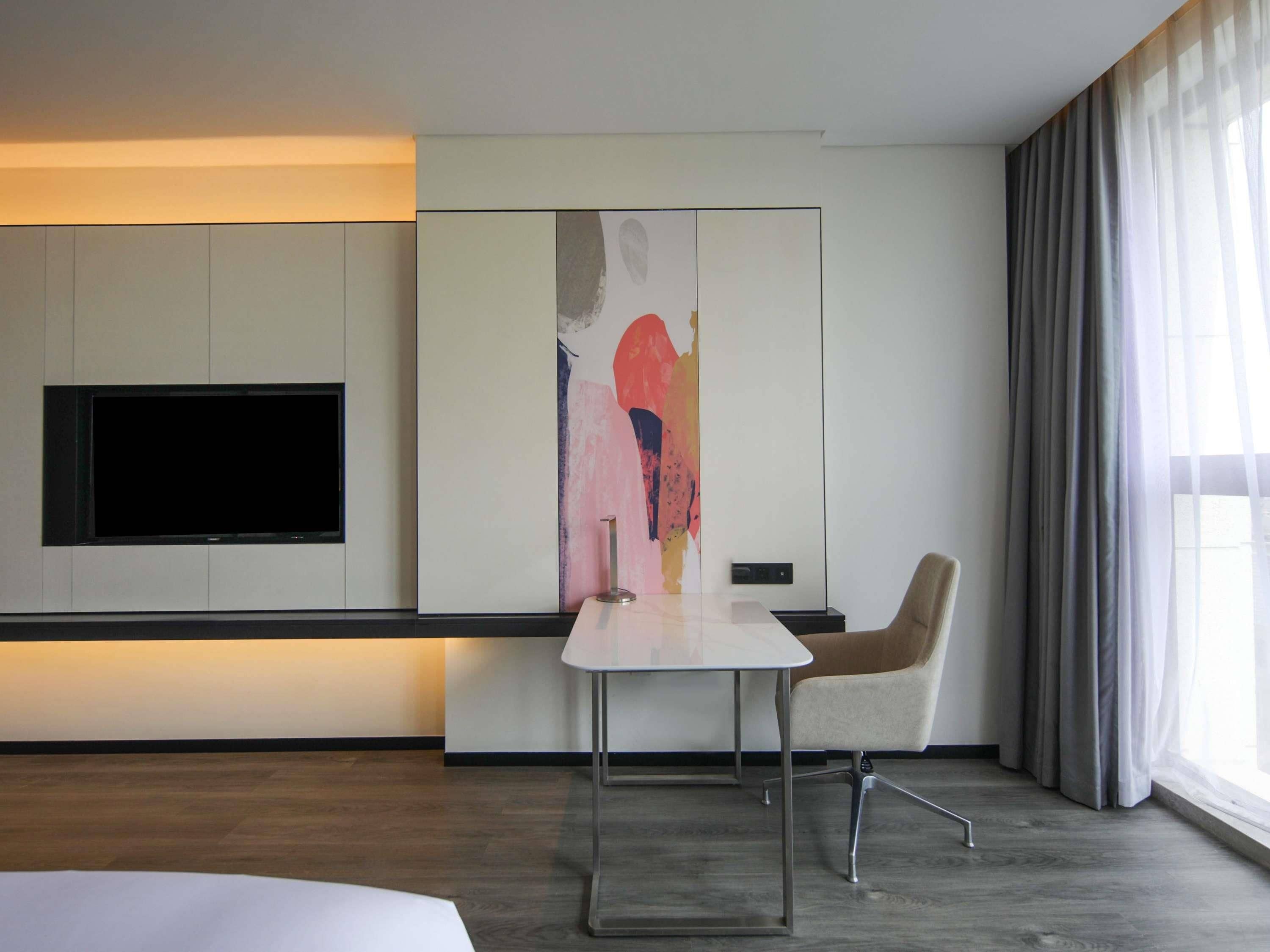 A modern hotel room with a flat-screen TV mounted on the wall, a minimalist table, a comfortable armchair, and a large window with curtains.
