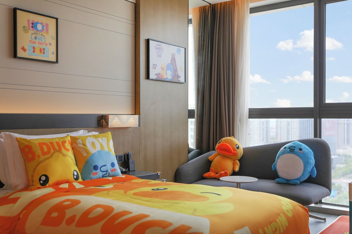 A hotel room with a bed featuring B. Duck pillows, a plush duck toy, and a framed picture on the wall.