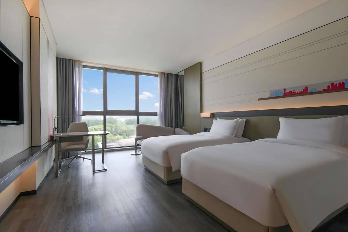 A hotel room with two beds, a desk, a chair, and a large window offering a city view.