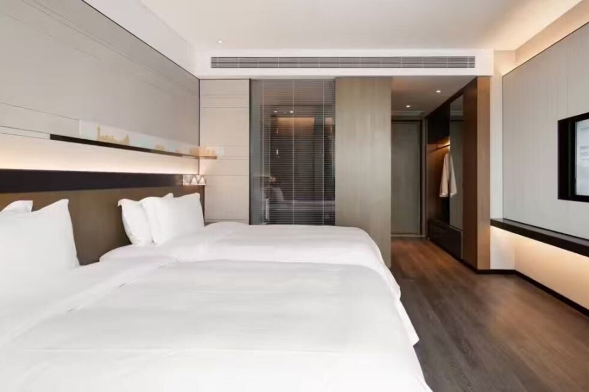 A modern hotel room with two beds, a large flat-screen TV, and a view of the cityscape through the window.