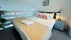 A neatly made bed with a white comforter and two rainbow pillows, a bedside lamp, and a small nightstand.