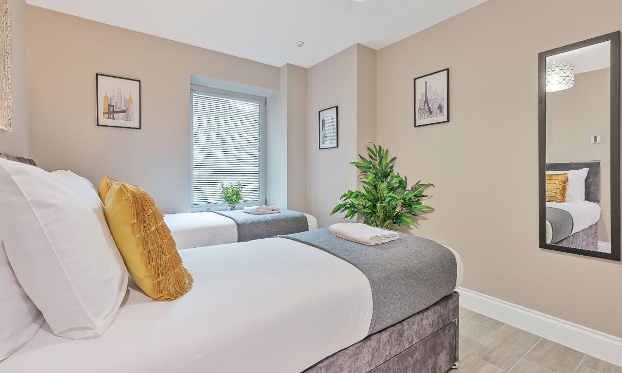 A bedroom with a bed, bedside tables, a window with blinds, a potted plant, and framed pictures on the walls.