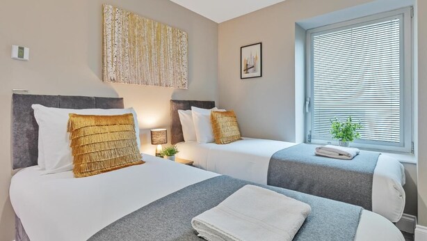 Habitación de hotel con dos camas, un ventanal con persianas, una obra de arte en la pared y una mesita de noche con lámpara.