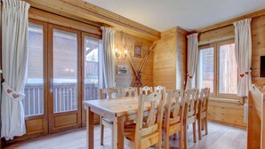 Ein Esszimmer mit Holzboden, einem großen Tisch und Stühlen, Holzvertäfelung und großen Fenstern mit Vorhängen.