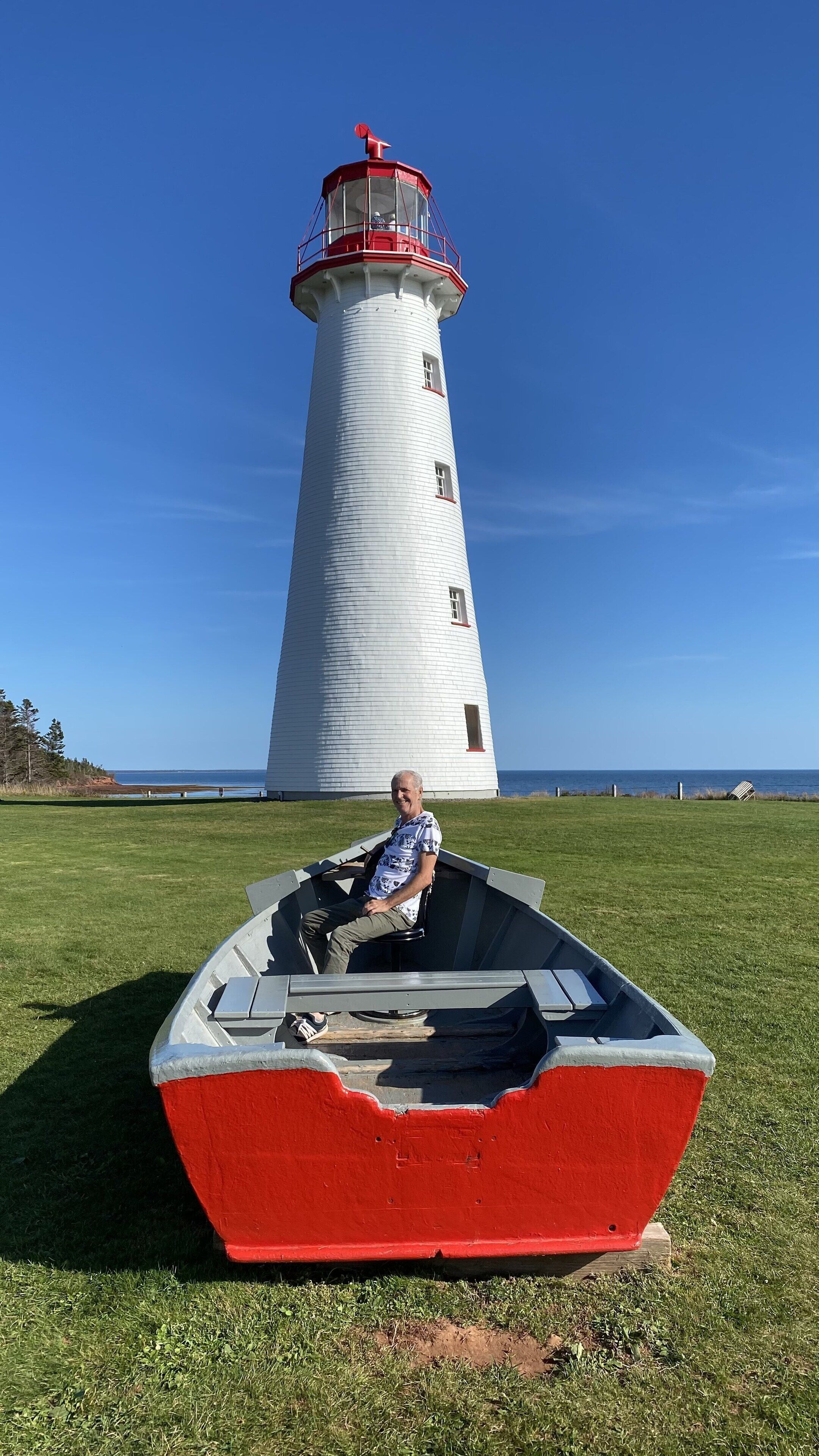 A person in a small boat with a red bottom and white top, positioned on a grassy field with a white lighthouse in the background.