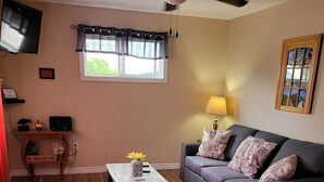 Un salon doté d’un plancher en bois, d’un ventilateur de plafond, d’un canapé gris agrémenté de coussins à motifs, d’un fauteuil rouge, d’une table basse en bois et d’un téléviseur fixé au mur.