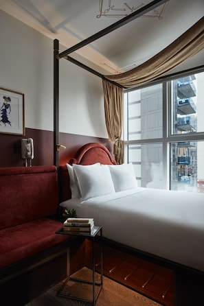 A hotel room with a red upholstered sofa, a bed with white linens, a small table with books, and a view of the city through large windows. - CIVILIAN Hotel (New York)