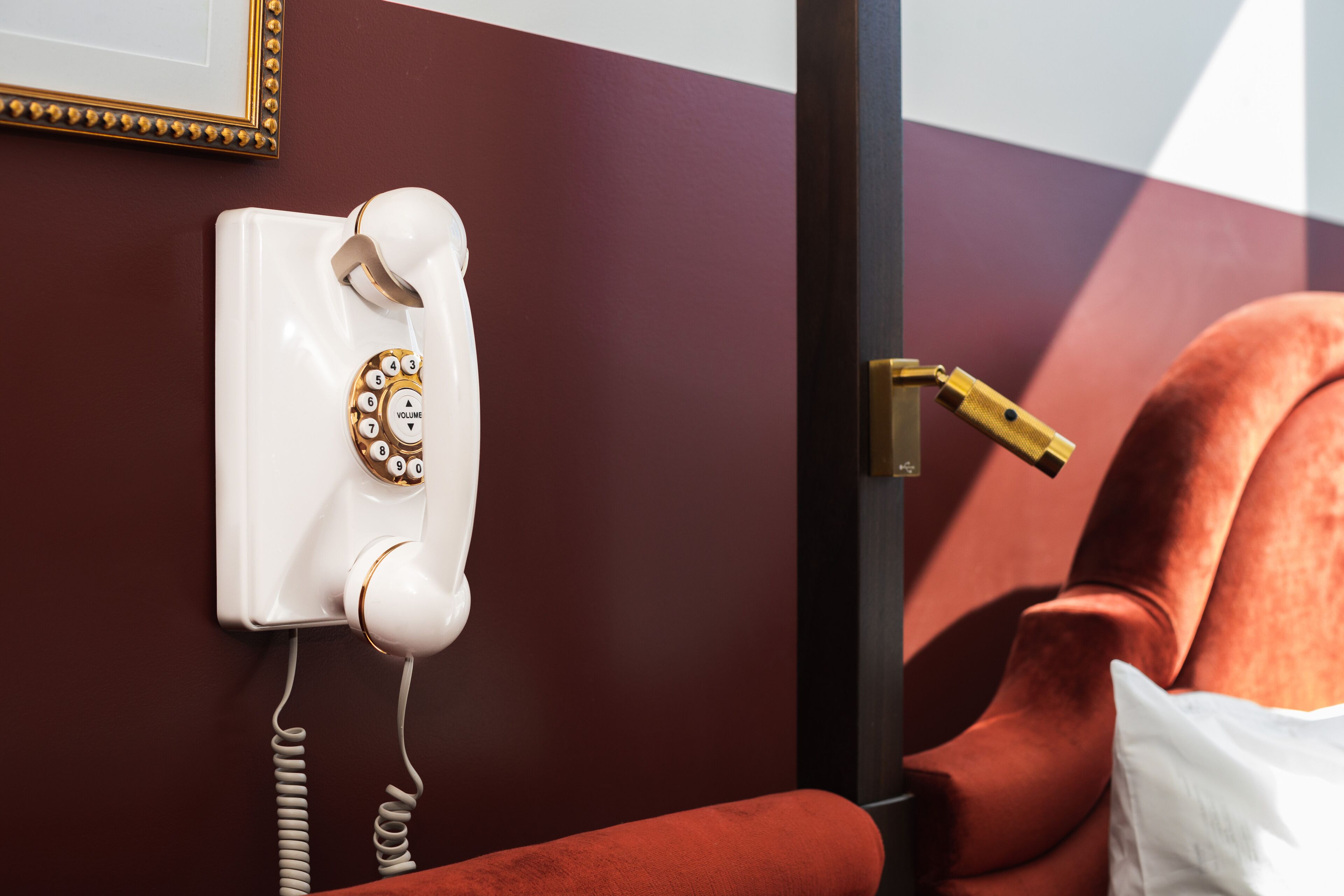 Ein altmodisches weißes Wählscheibentelefon an der Wand, eingefasst in Gold, ein roter Samtesselstuhl und ein weißes Kissen auf einem schwarzen Stuhl.