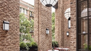 A brick-walled outdoor patio with round tables and red chairs, potted plants, and hanging lanterns.