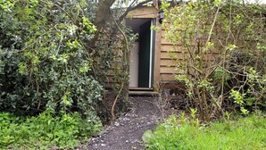 Un estrecho sendero de grava que conduce a una estructura de madera con una puerta verde, rodeada de abundante vegetación.
