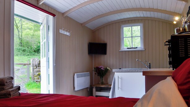 Un dormitorio con colcha roja, una pequeña kitchenette con lavamanos y estufa, un televisor montado en la pared y una ventana con vistas a la vegetación.