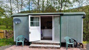Un cobertizo metálico de una sola planta con una puerta blanca y un pequeño porche. En su interior, hay una cocina con armarios y una encimera. Dos sillas de plástico verde están colocadas en el porche.