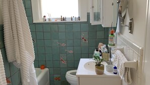 Kylpyhuone, jossa on valkoinen kylpyamme, wc, pesuallas, jossa on hygieniatuotteita, ja ikkuna.