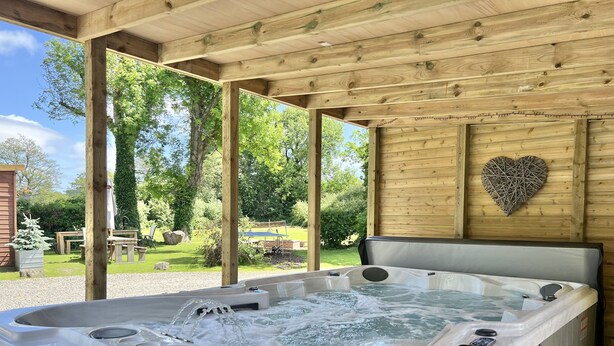 A hot tub with multiple jets, surrounded by a wooden deck and a heart-shaped wall decoration. - Beautiful Large Country Barn Conversion Hot Tub, Pool, BBQ Hut, Large Grounds (Lampeter)