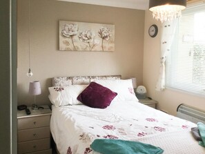 A bedroom with a bed, a nightstand, a lamp, a clock, and a window with blinds.