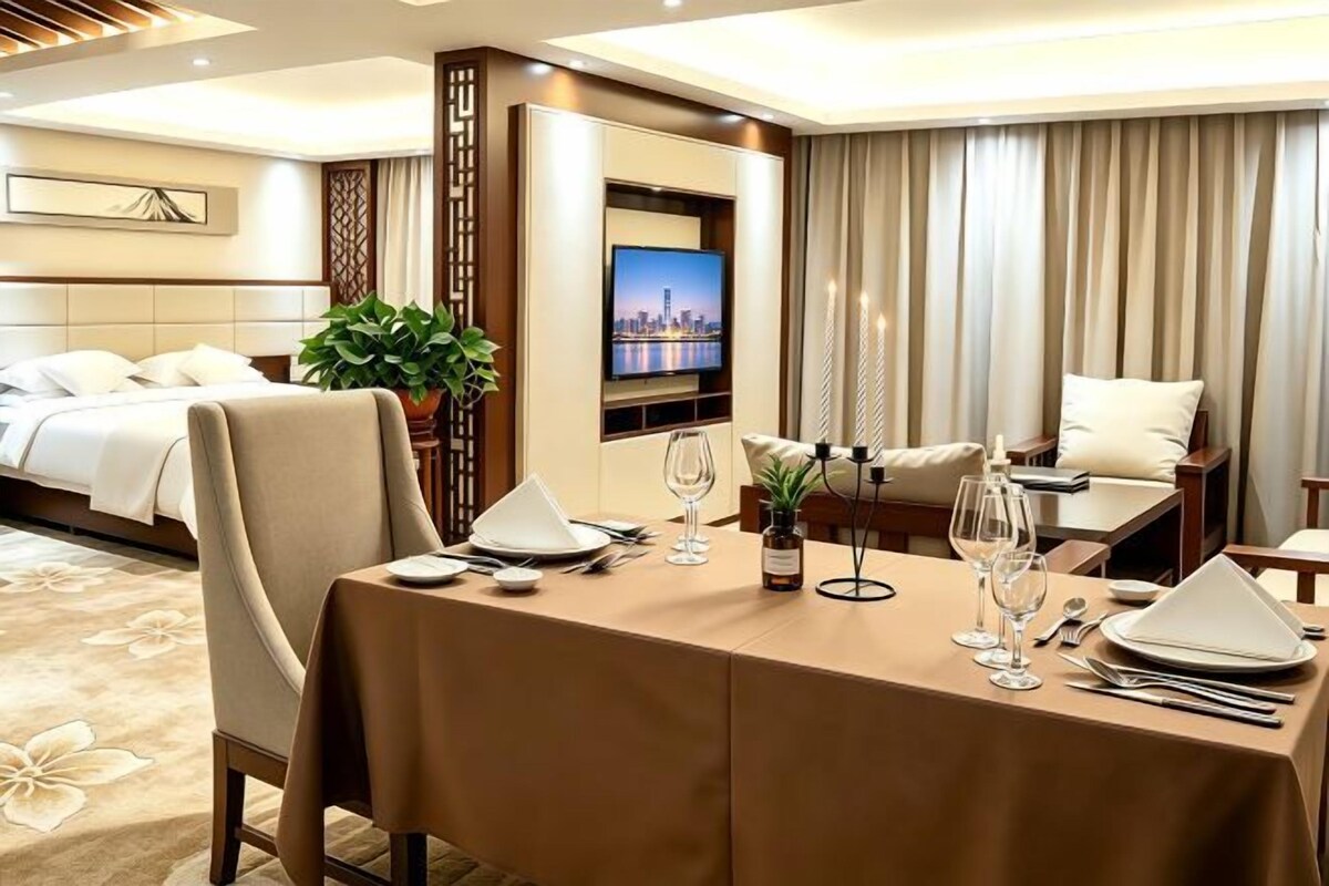 A hotel room with a bed, a dining table set for two, a TV, and a view of the cityscape.