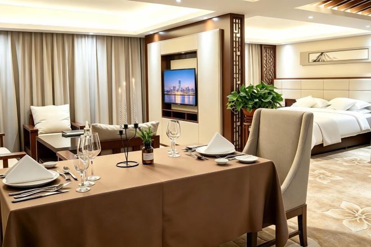 A hotel room with a dining table set for two, a bed with white linens, a television, and a cityscape view.