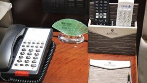 Un bureau dans une chambre d’hôtel, avec un téléphone, un ventilateur vert, une boîte de télécommandes et un stylo.