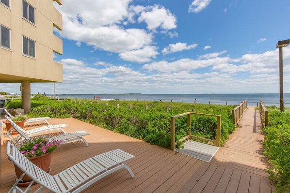 A wooden deck with lounge chairs and a potted plant, overlooking a beach and ocean.