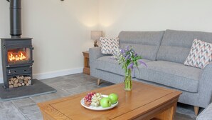 Een woonkamer met een houtkachel, een grijze bank, een houten salontafel met fruit en wandlampen.